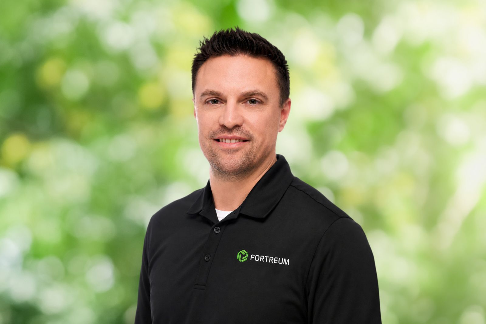 Smiling man with short dark hair wearing a black Fortreum polo shirt against a blurred green foliage background.