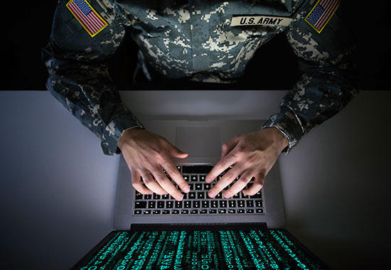 U.S. Army soldier in camouflage uniform typing on a laptop displaying glowing green code on the screen.