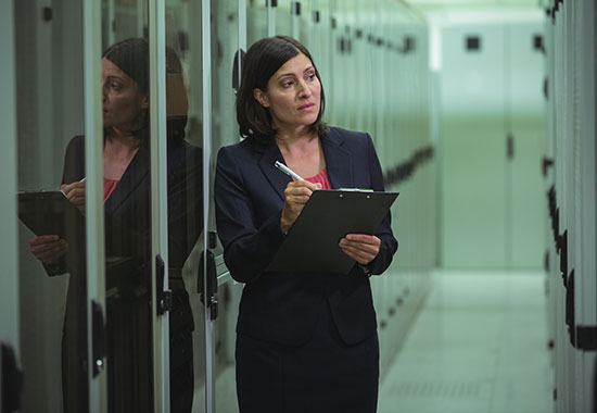 Professional woman in business attire holding a clipboard while inspecting equipment in a data center server room.