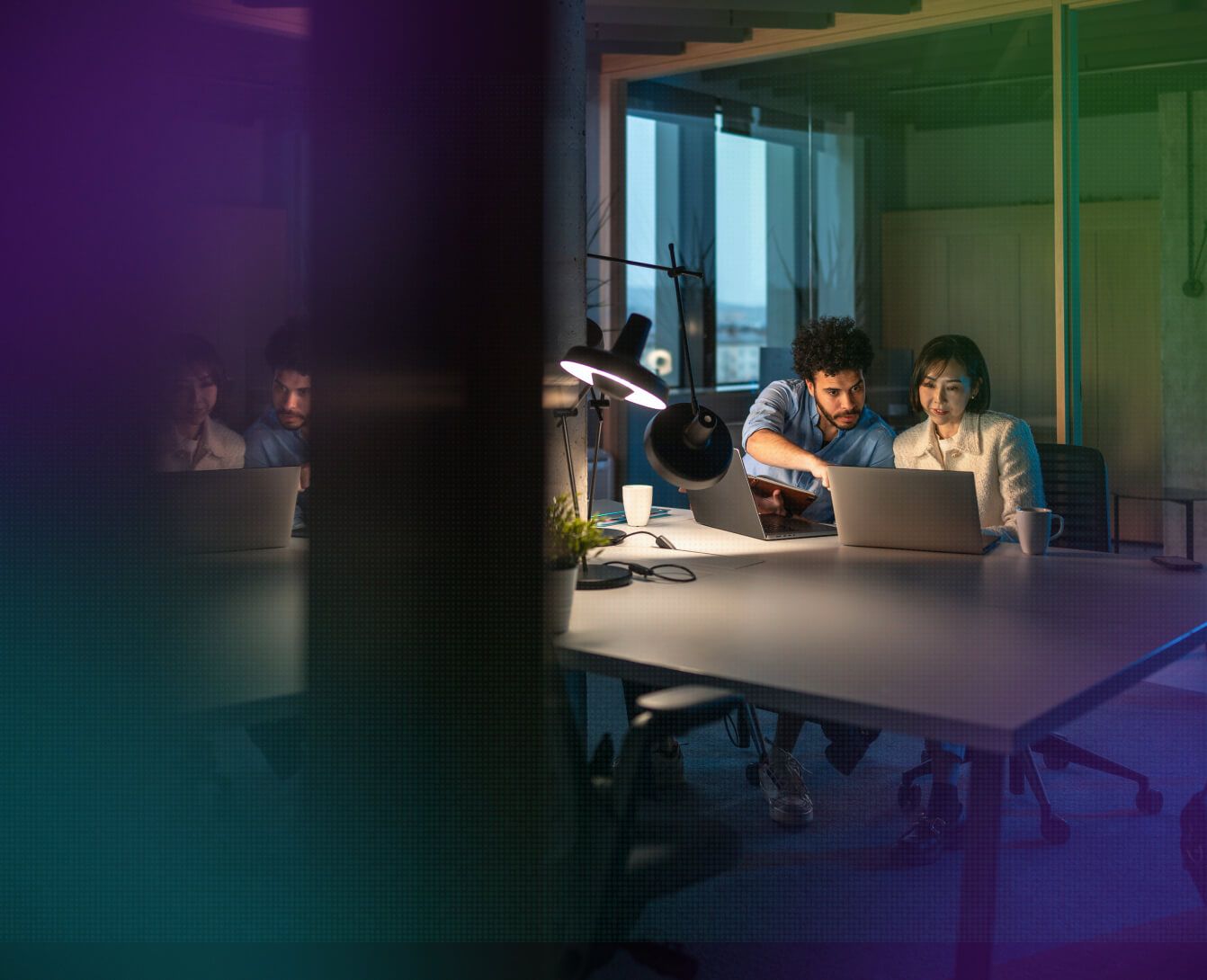 Colleagues collaborate on laptops around a desk lamp in a dimly lit modern office at night.