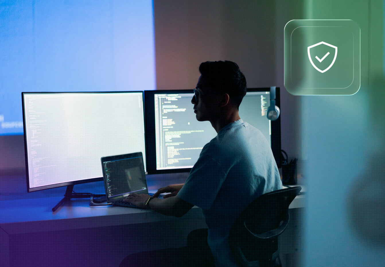 Cybersecurity professional working at a multi-monitor workstation with code displayed, alongside a security shield icon.