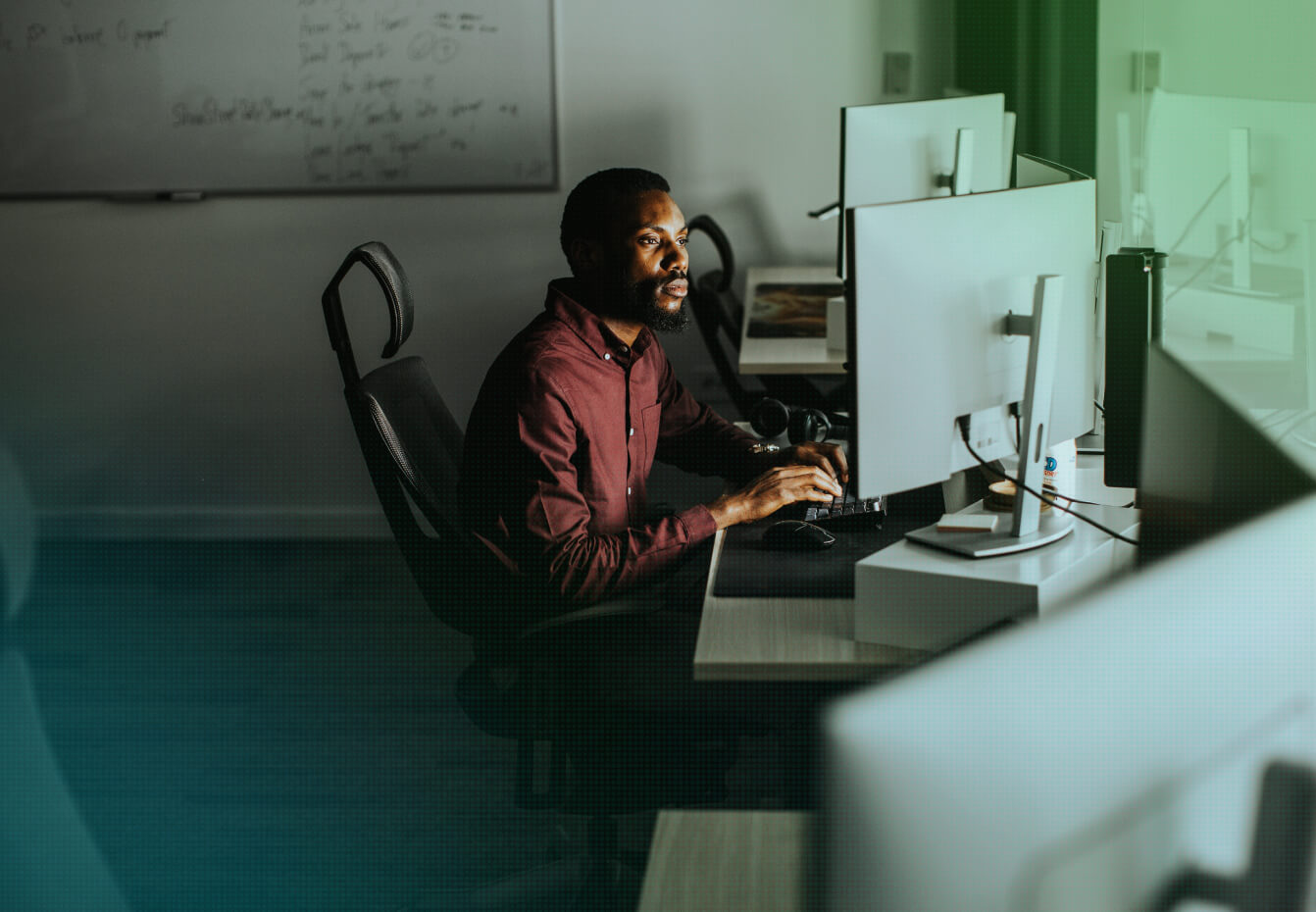 Focused man in a maroon shirt working at a dual-monitor computer workstation in a dimly lit office with a whiteboard.