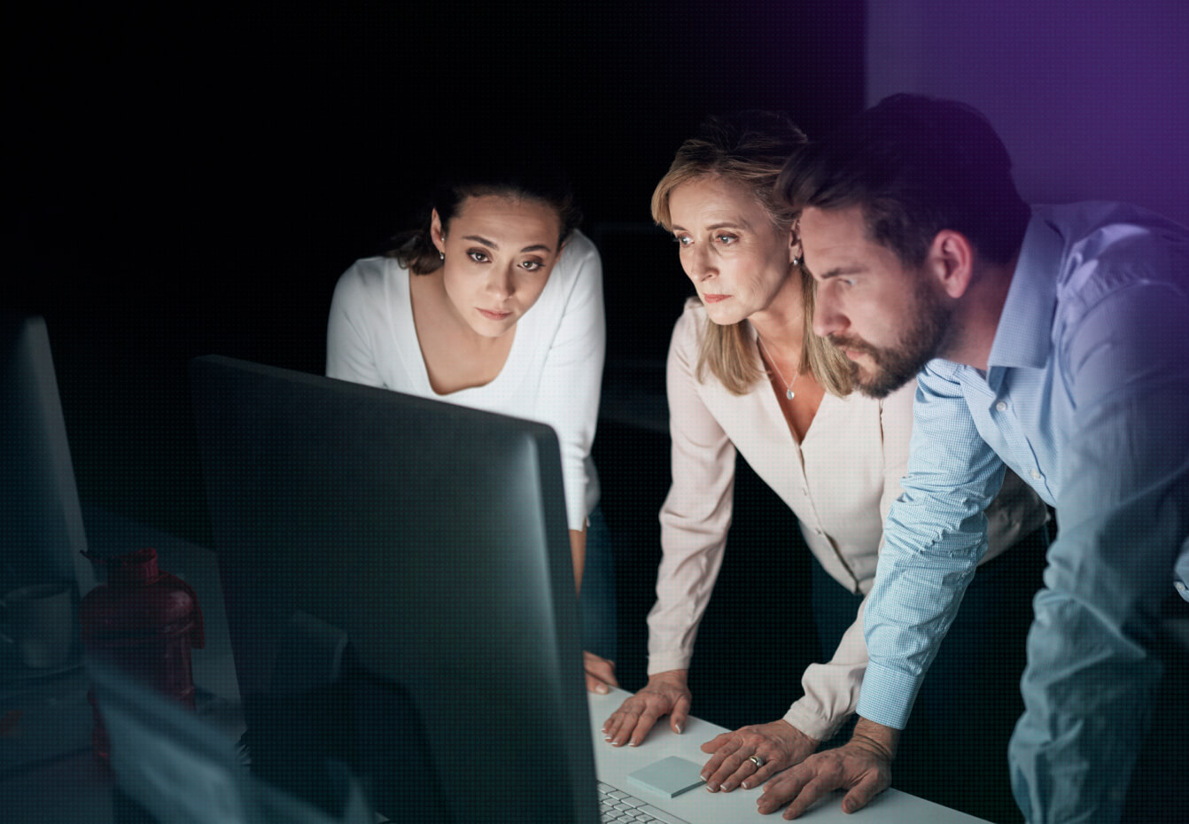 Fortreum-CMMC-Mission-Our-C3PAO-Card2 Three professionals focused intently on a computer monitor in a dimly lit office, collaborating on a cybersecurity task.