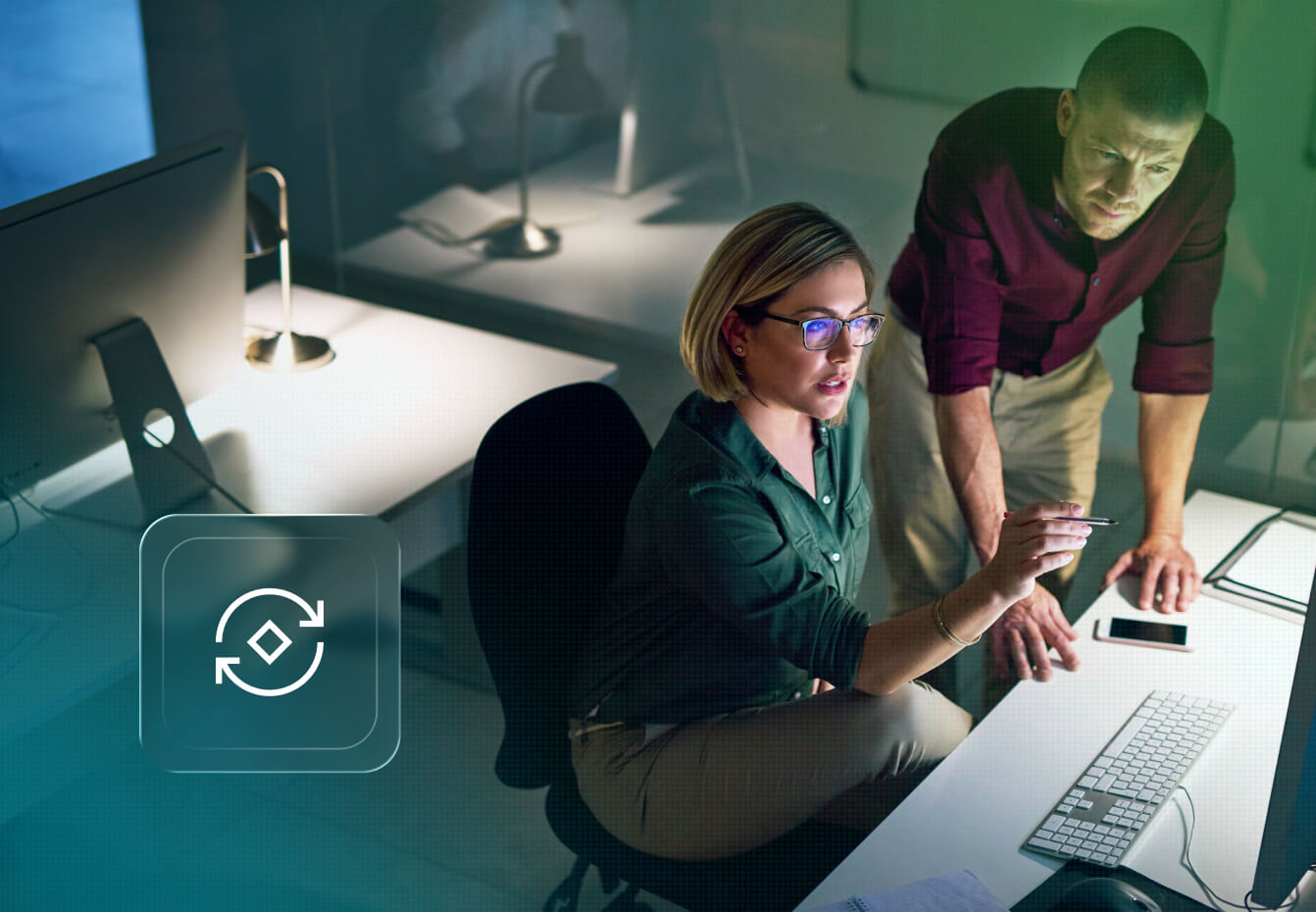 Two cybersecurity professionals collaborate at a computer workstation in a dimly lit office, reviewing data together.