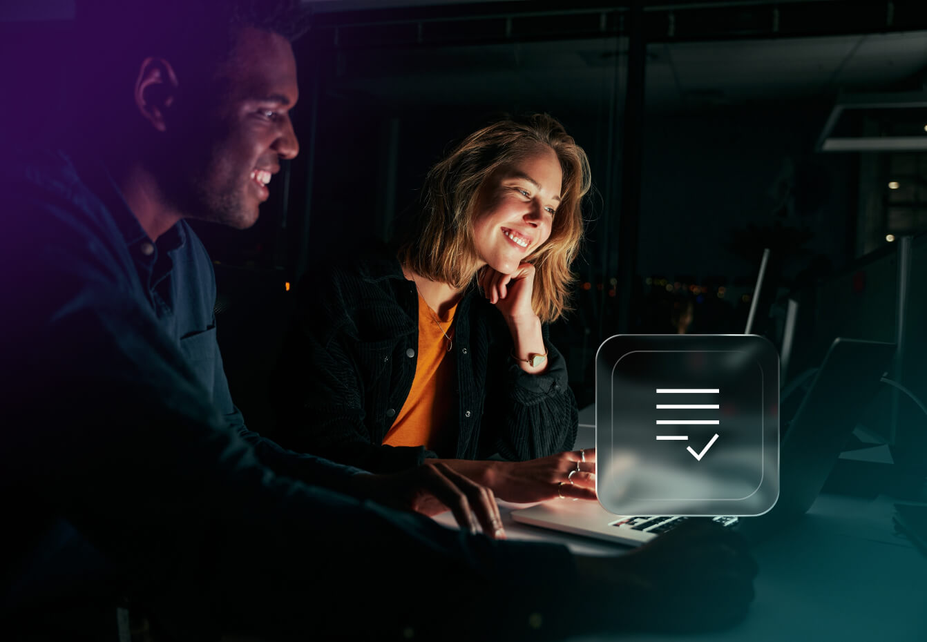 Two smiling colleagues collaborate at a laptop in a dimly lit office, with a checklist icon overlay representing assessment
