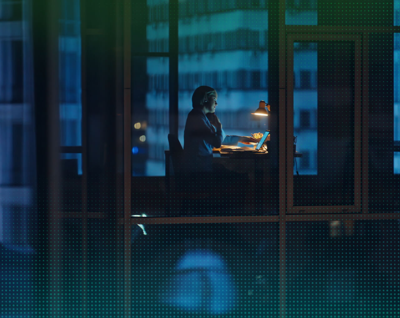 Person working late at a desk with a lit lamp and laptop in a high-rise office, viewed through windows at night.