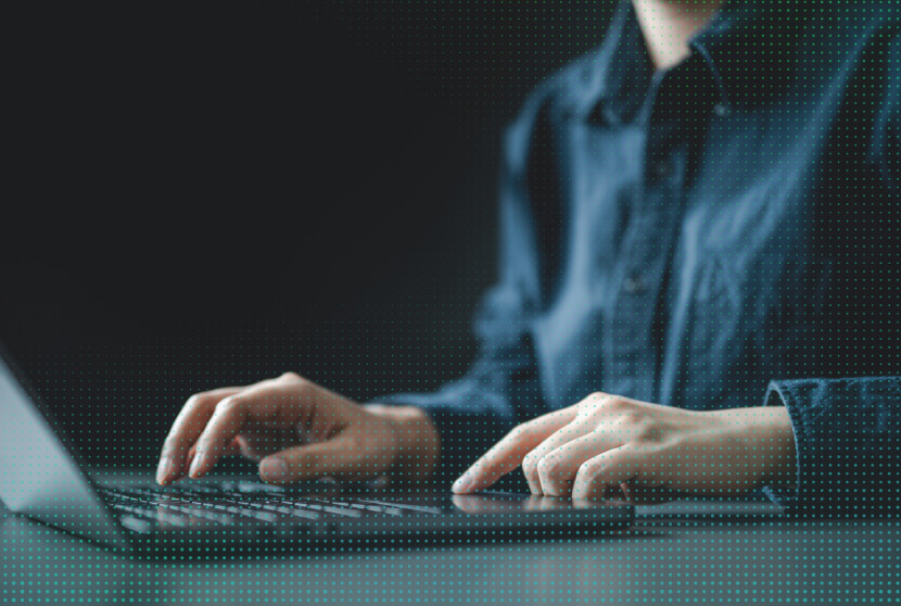 Person in a denim shirt typing on a laptop keyboard against a dark background with a digital dot overlay.