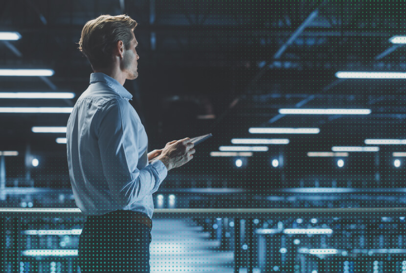 Man in blue shirt holding a tablet while standing in a modern data center with illuminated server racks.