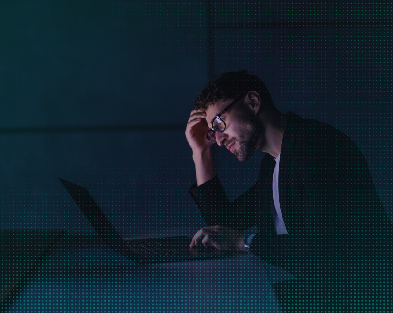 Stressed man with glasses working on a laptop in a dimly lit room, holding his head in frustration.
