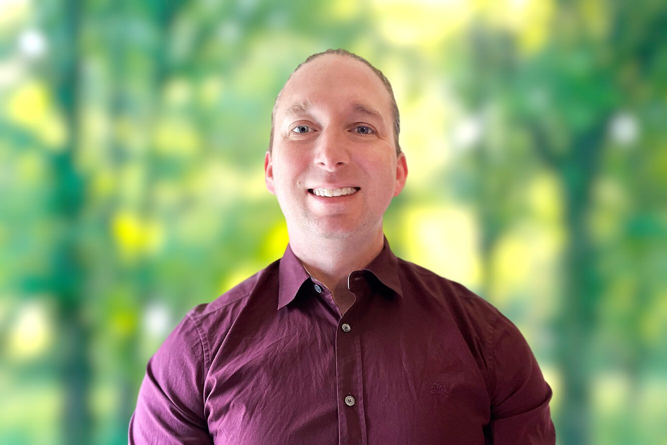 Smiling man in a maroon button-down shirt poses against a blurred green and yellow nature background.