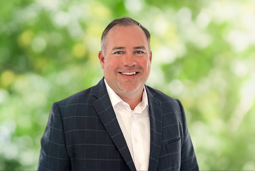 Smiling man in a dark checkered blazer and white shirt poses against a blurred green foliage background.