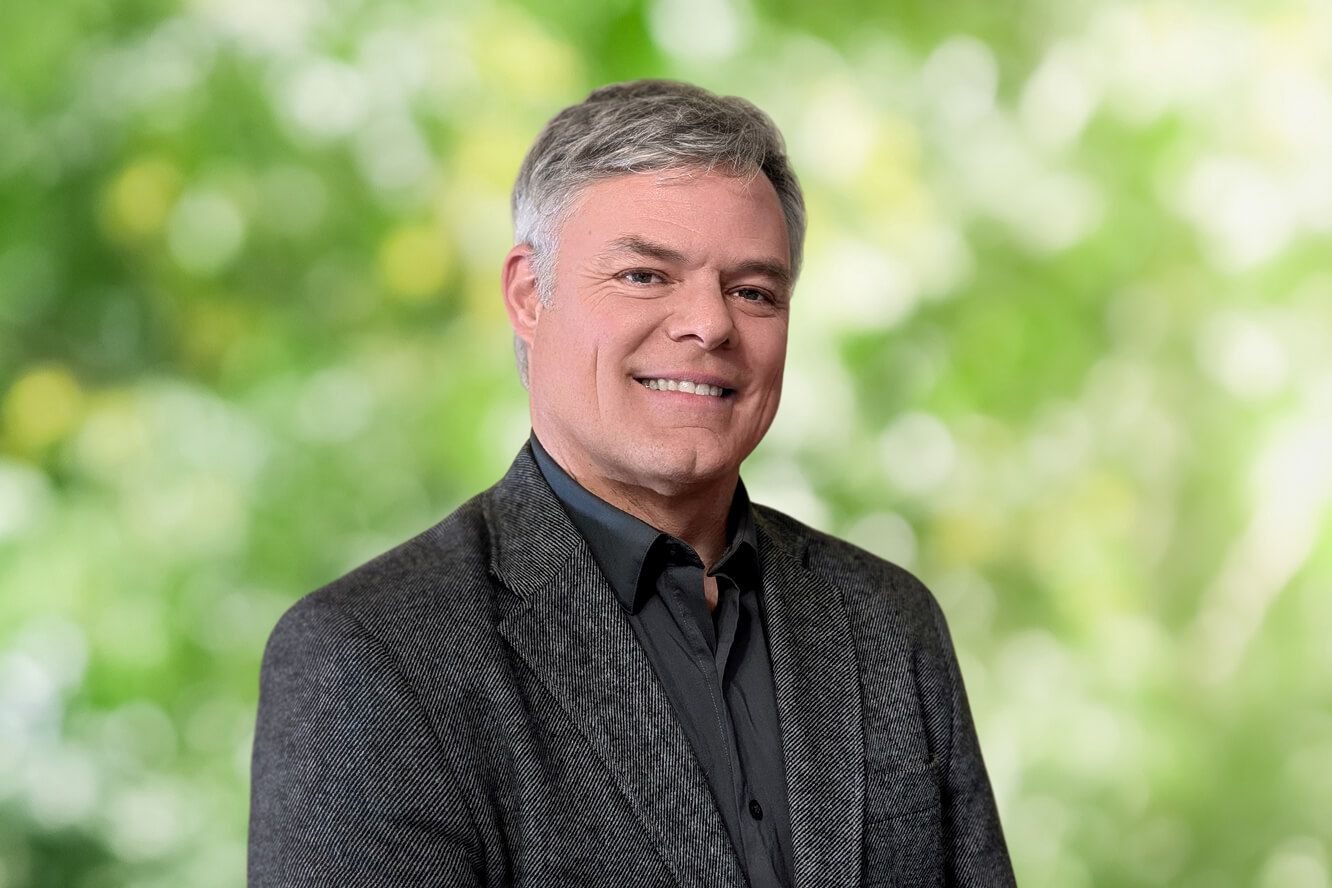 Smiling man with gray hair wearing a charcoal blazer and black shirt against a blurred green foliage background.