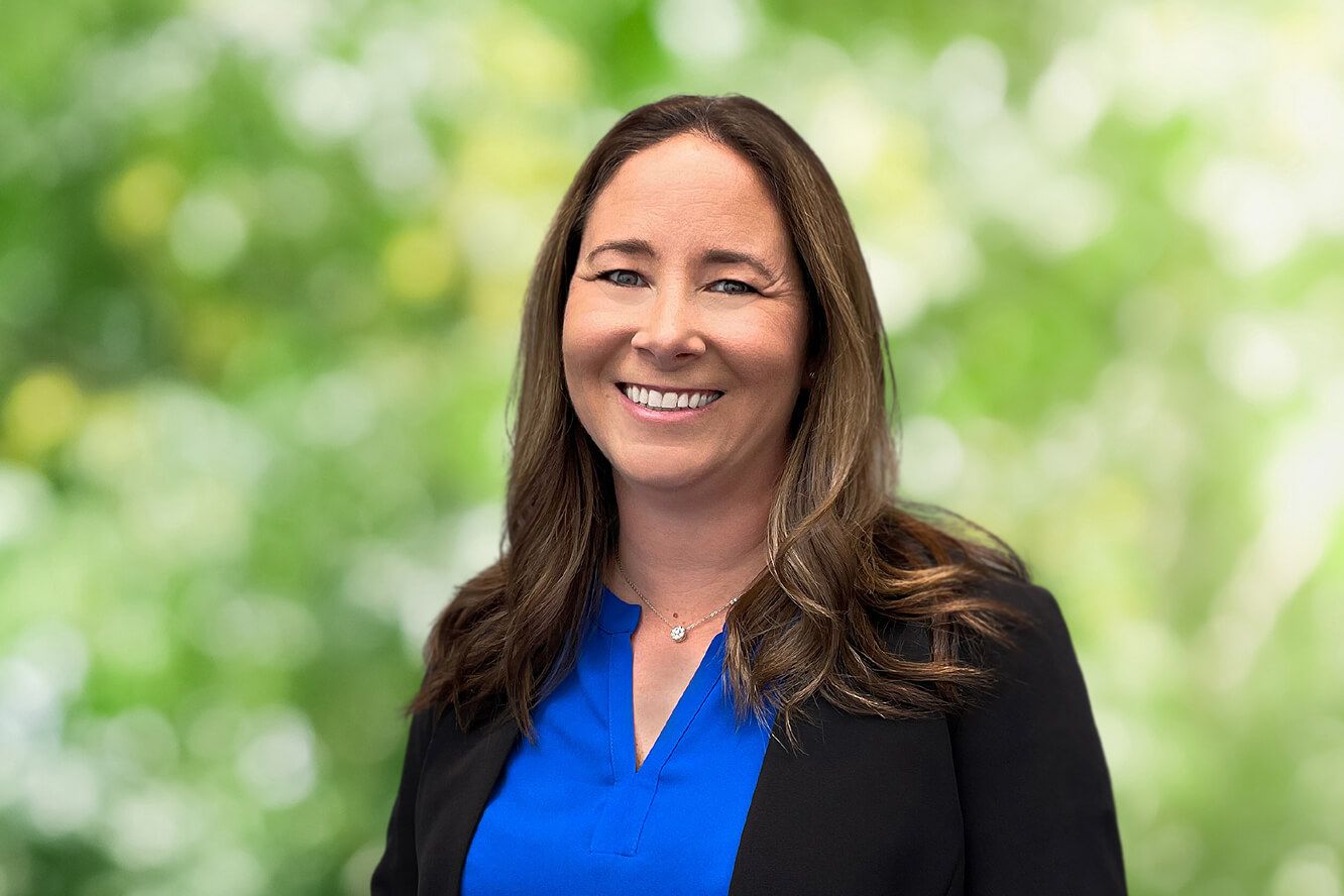 Smiling professional woman with long brown hair wearing a blue blouse and black blazer against a blurred green background.