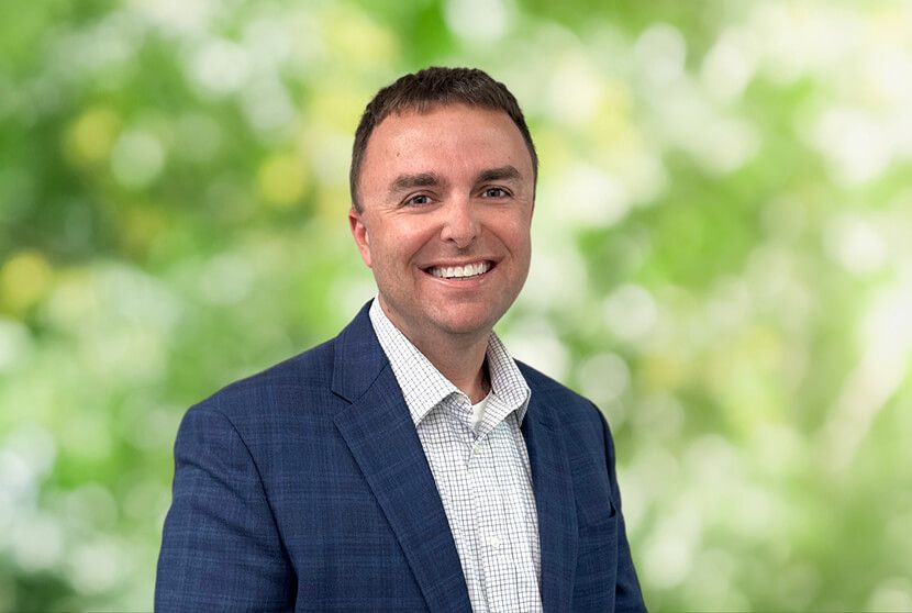 Michael Carter smiling in a navy blazer and checkered shirt against a blurred green foliage background.
