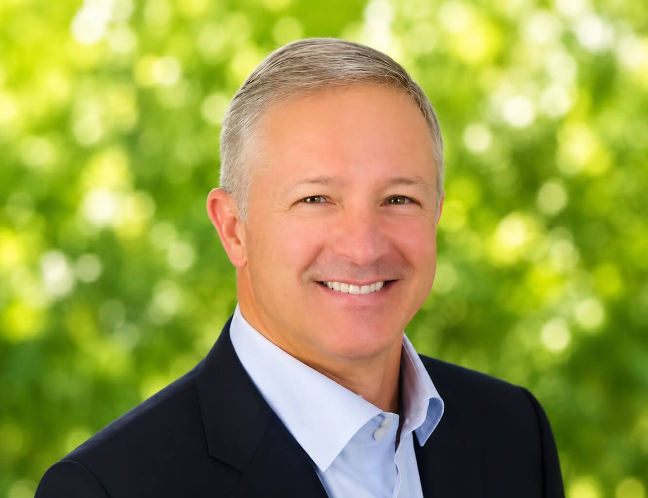 Smiling man with short gray hair wearing a light blue shirt and dark navy blazer against a blurred green foliage background.