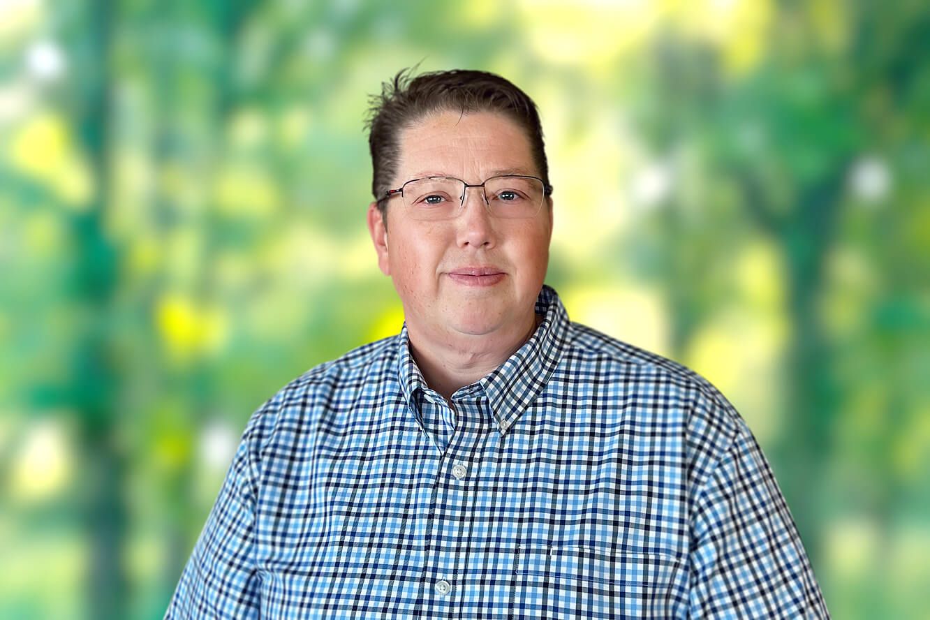 Rich Fleeman wearing glasses and a blue checkered shirt against a blurred green foliage background.