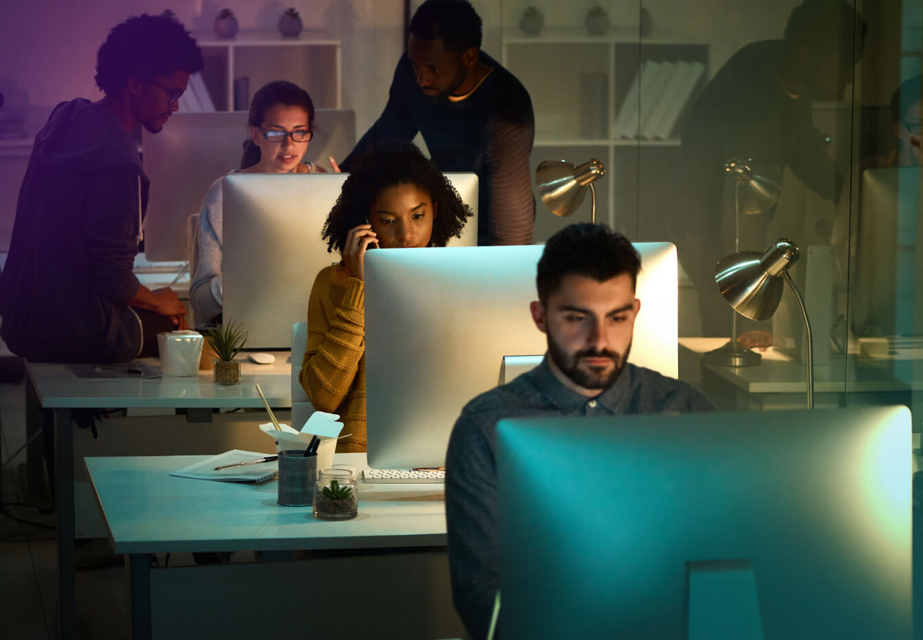 Diverse team of professionals working late at computer workstations in a dimly lit modern office environment.