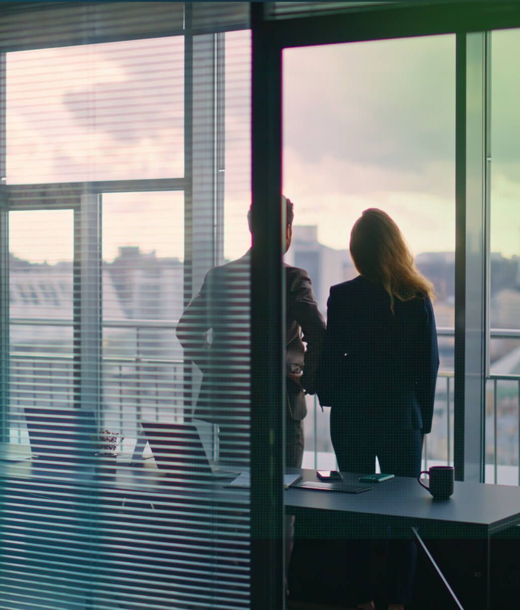 Two business professionals stand silhouetted by a large office window overlooking a city skyline at dusk.