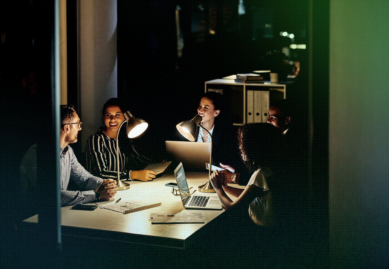 Four colleagues collaborate around a table with laptops and desk lamps during a late-night office work session.