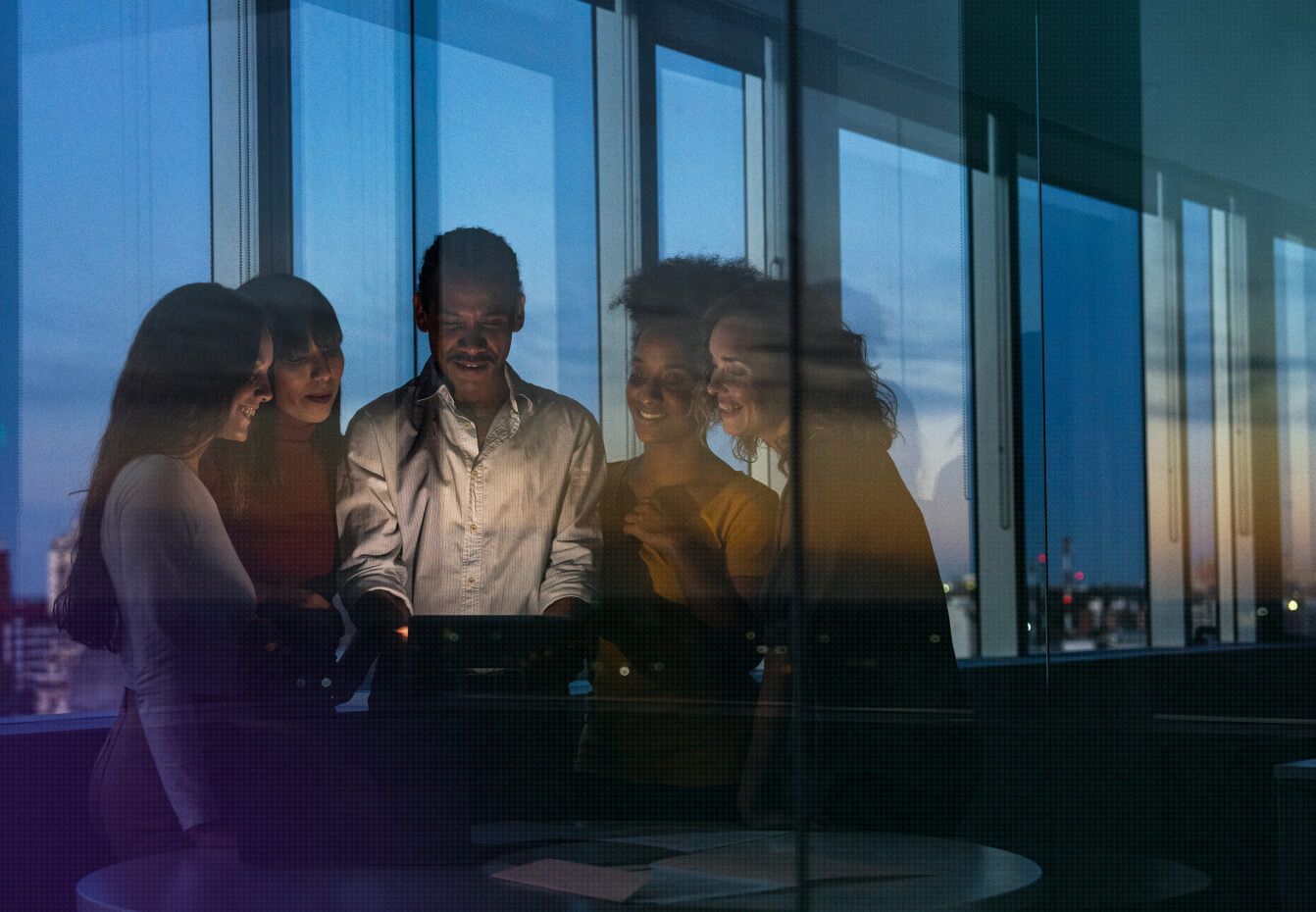 Diverse team of colleagues collaborating around a laptop in a modern office at dusk, viewed through glass windows.