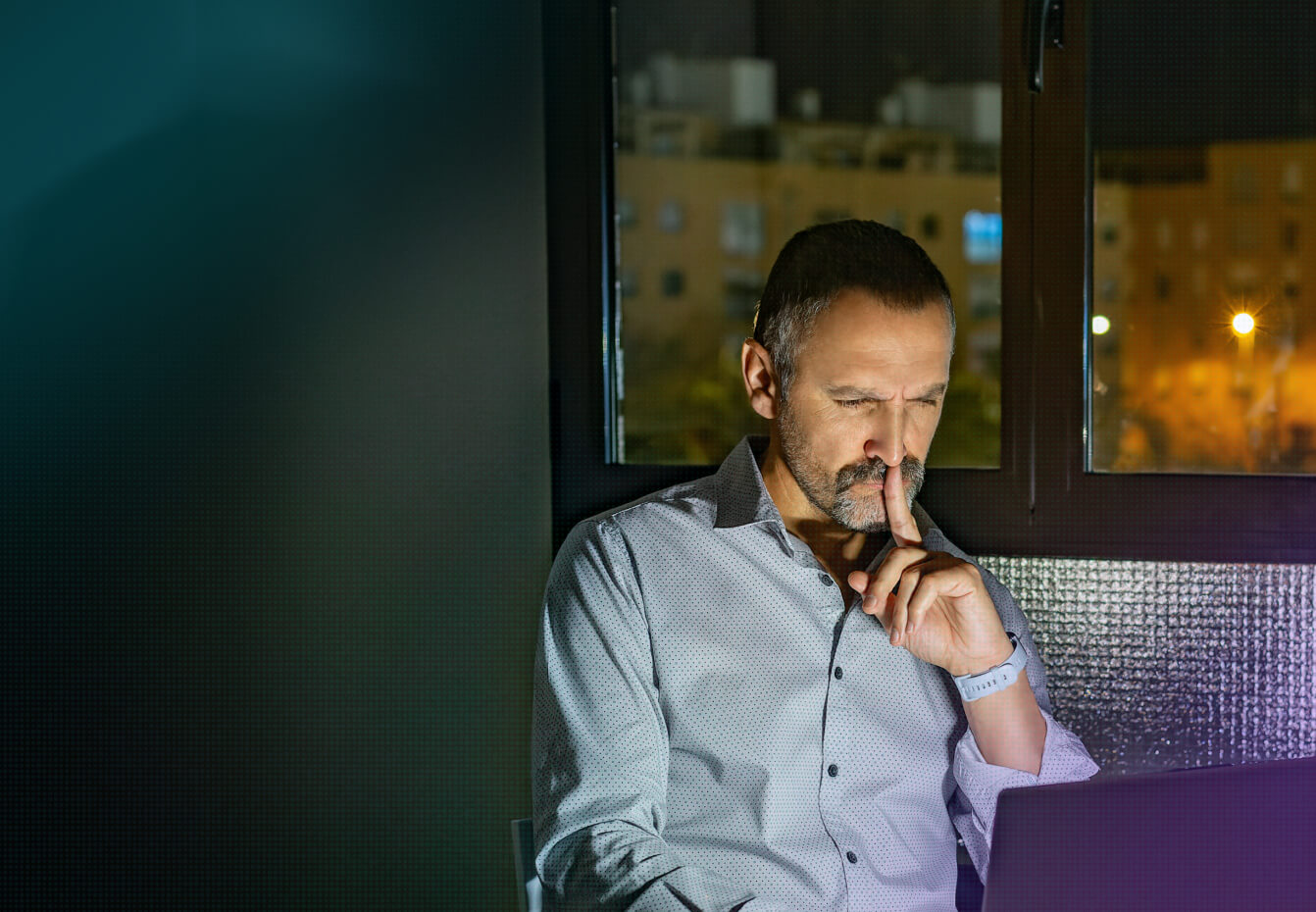 Thoughtful man in a gray shirt working on a laptop at night near a window with city lights in the background.