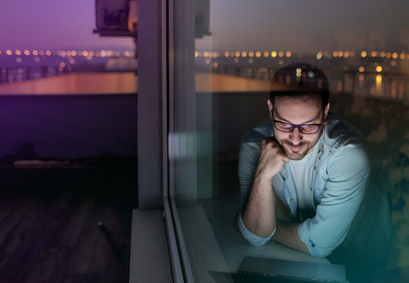 Man with glasses working on a laptop at night by a window overlooking a city skyline with bokeh lights.