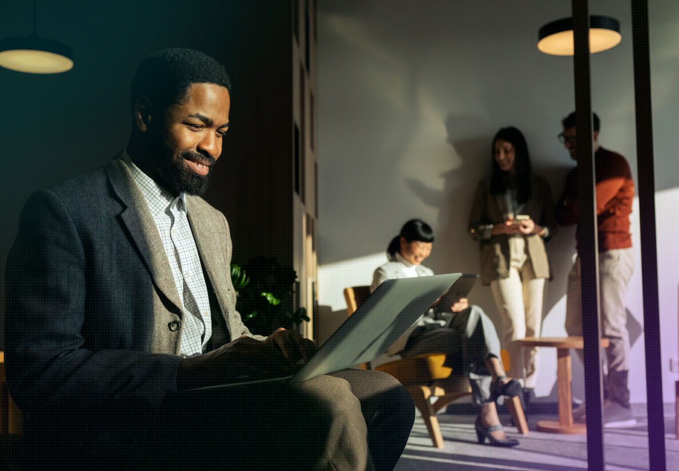 Bearded professional in a blazer smiles while working on a laptop as colleagues collaborate in the background office space.