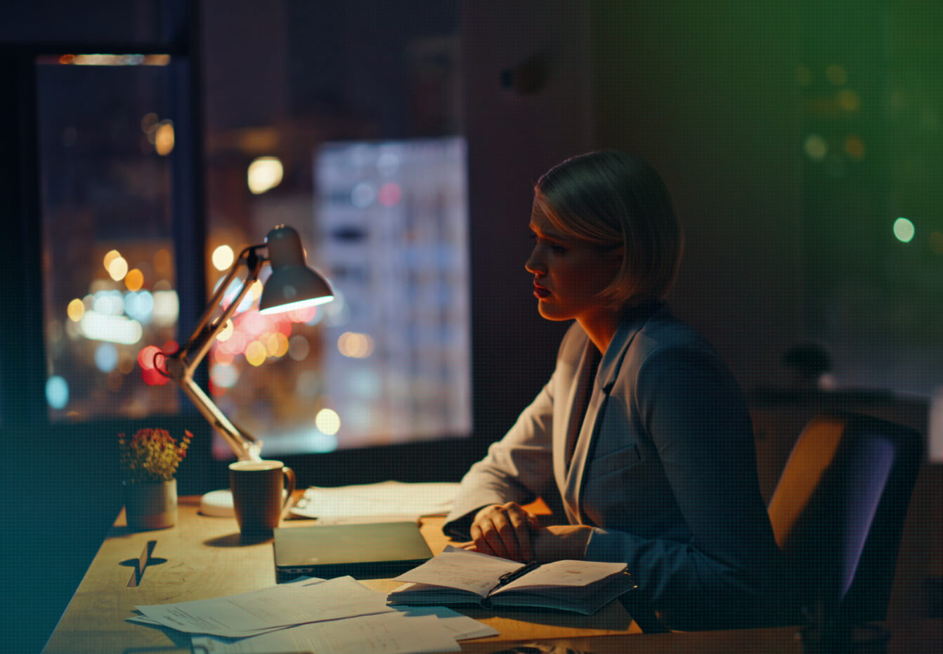 Woman in business attire works late at a desk illuminated by a lamp, with city lights glowing through the window behind her.