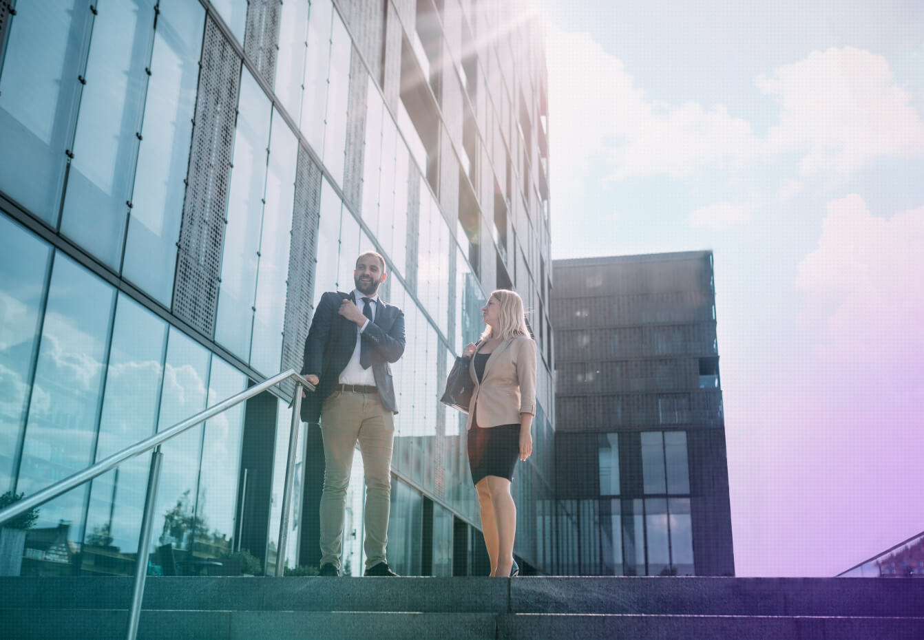 Two business professionals converse on outdoor steps outside a modern glass office building under bright sunlight.