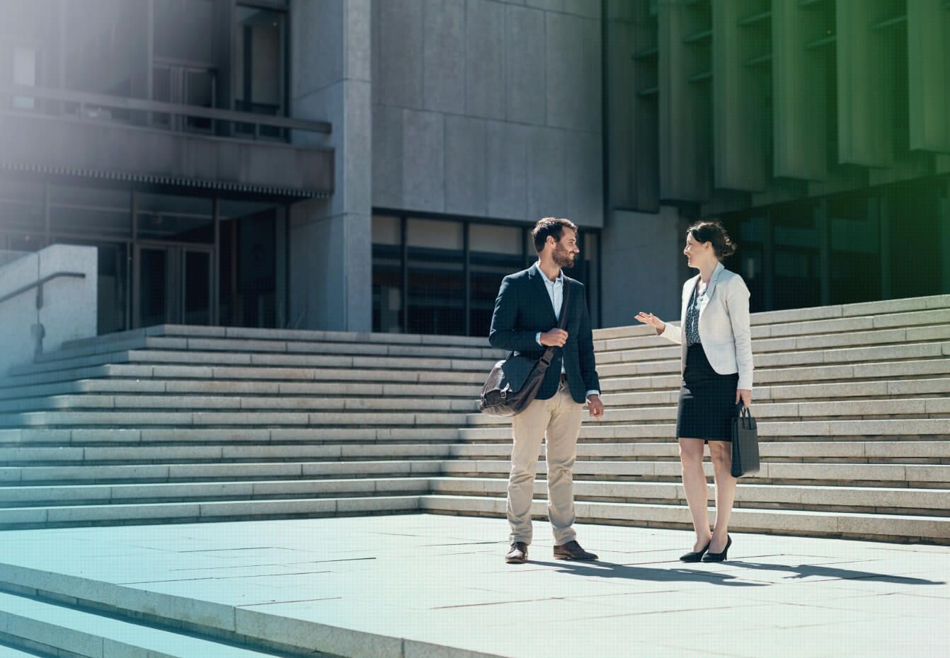 Two business professionals converse outside a modern office building near concrete steps, carrying a messenger bag and