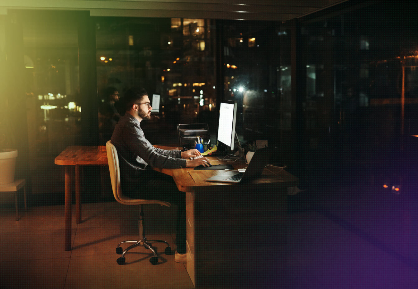 Man working late at night on a desktop computer and laptop in a darkened office with city lights visible through windows.