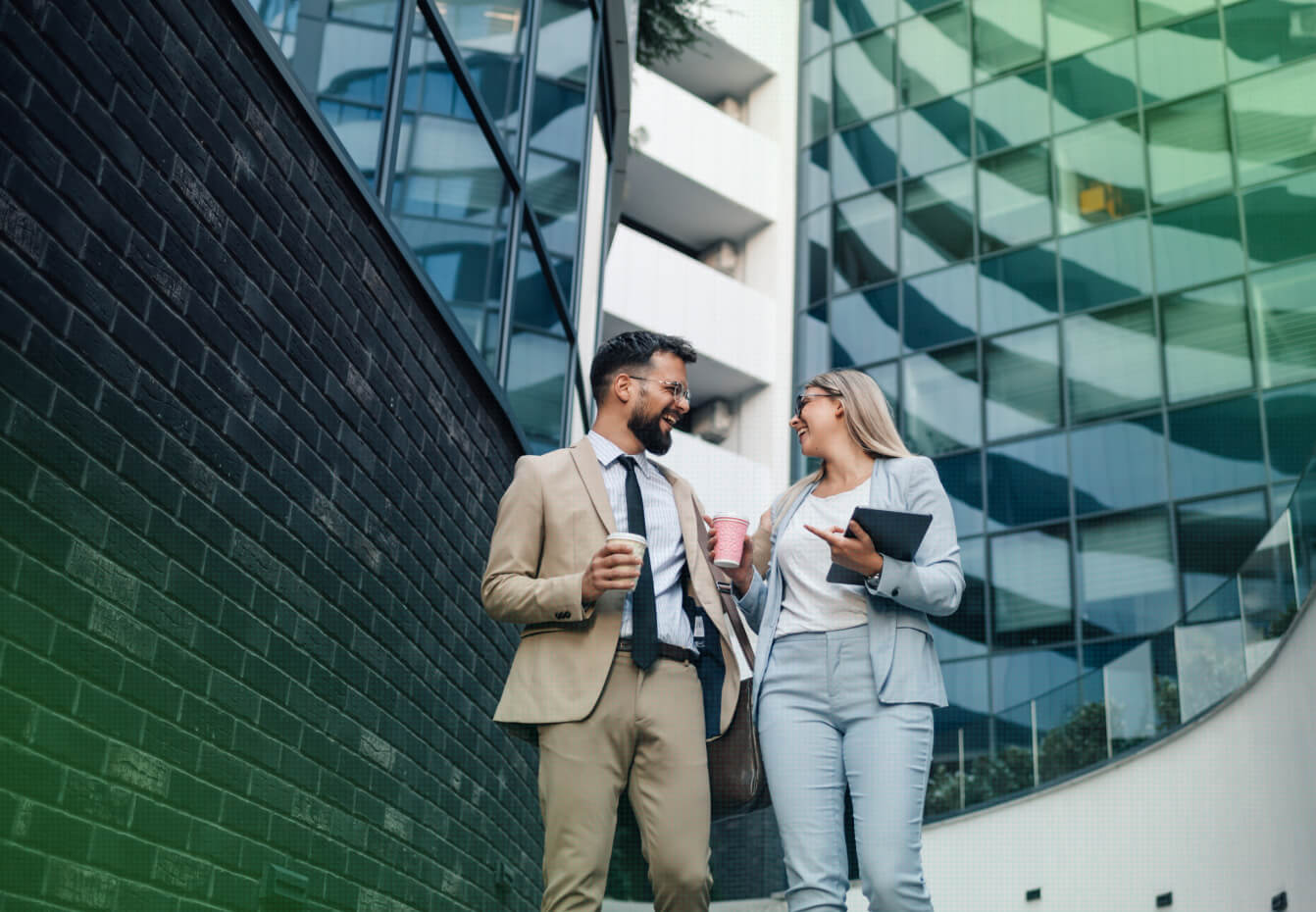 Two business professionals laugh together while holding coffee cups and a tablet outside a modern glass office building.