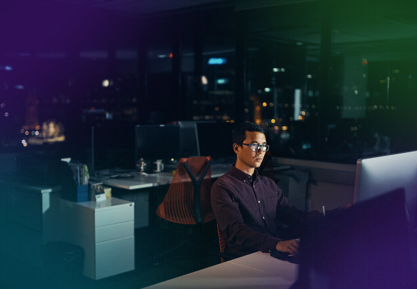 Man in glasses working late at a computer in a high-rise office with city lights visible through windows at night.