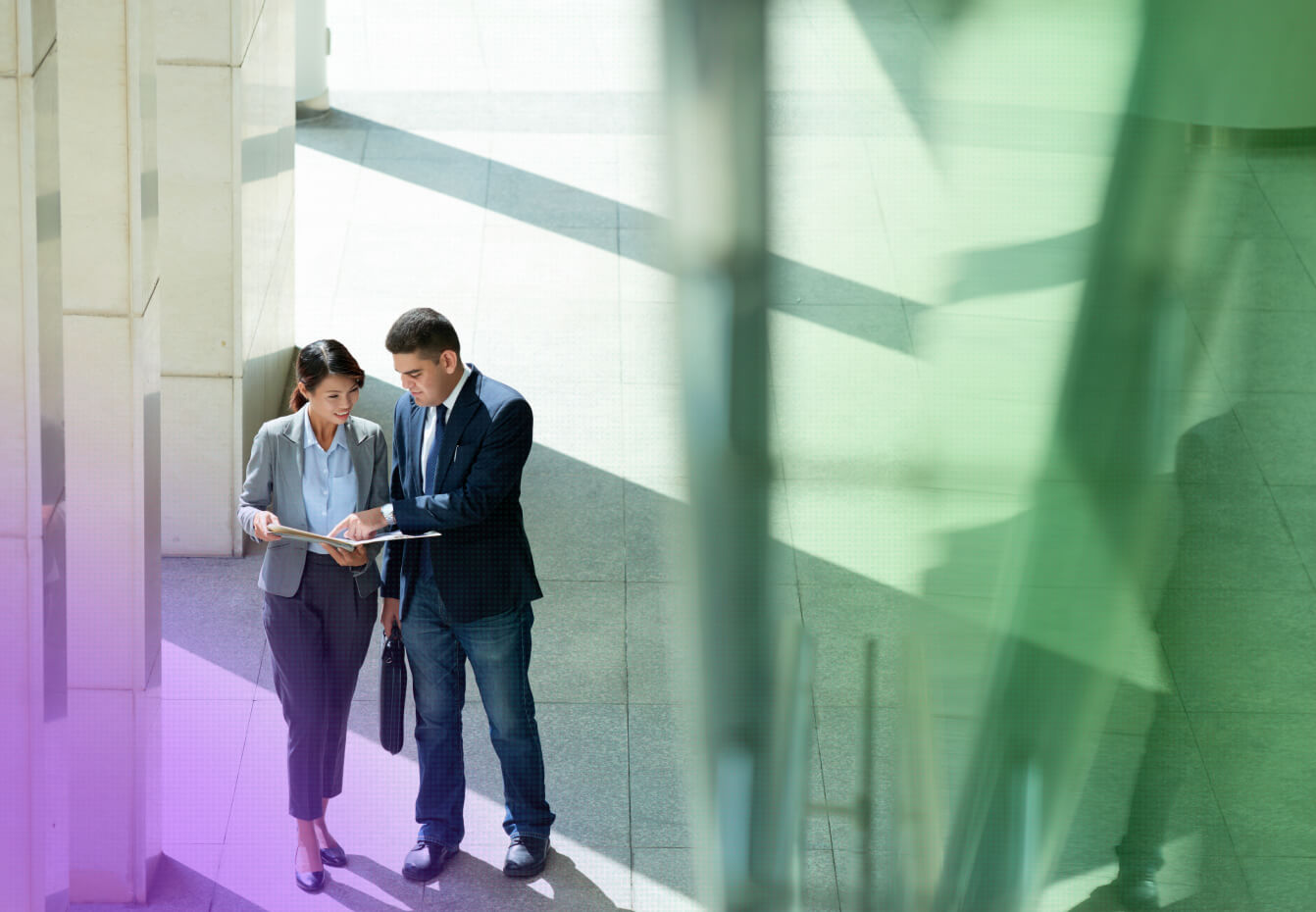 Two business professionals review documents together while standing in a sunlit modern office lobby.