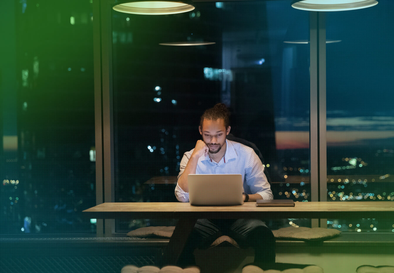 Man working on a laptop at night in a high-rise office with a city skyline visible through the window.