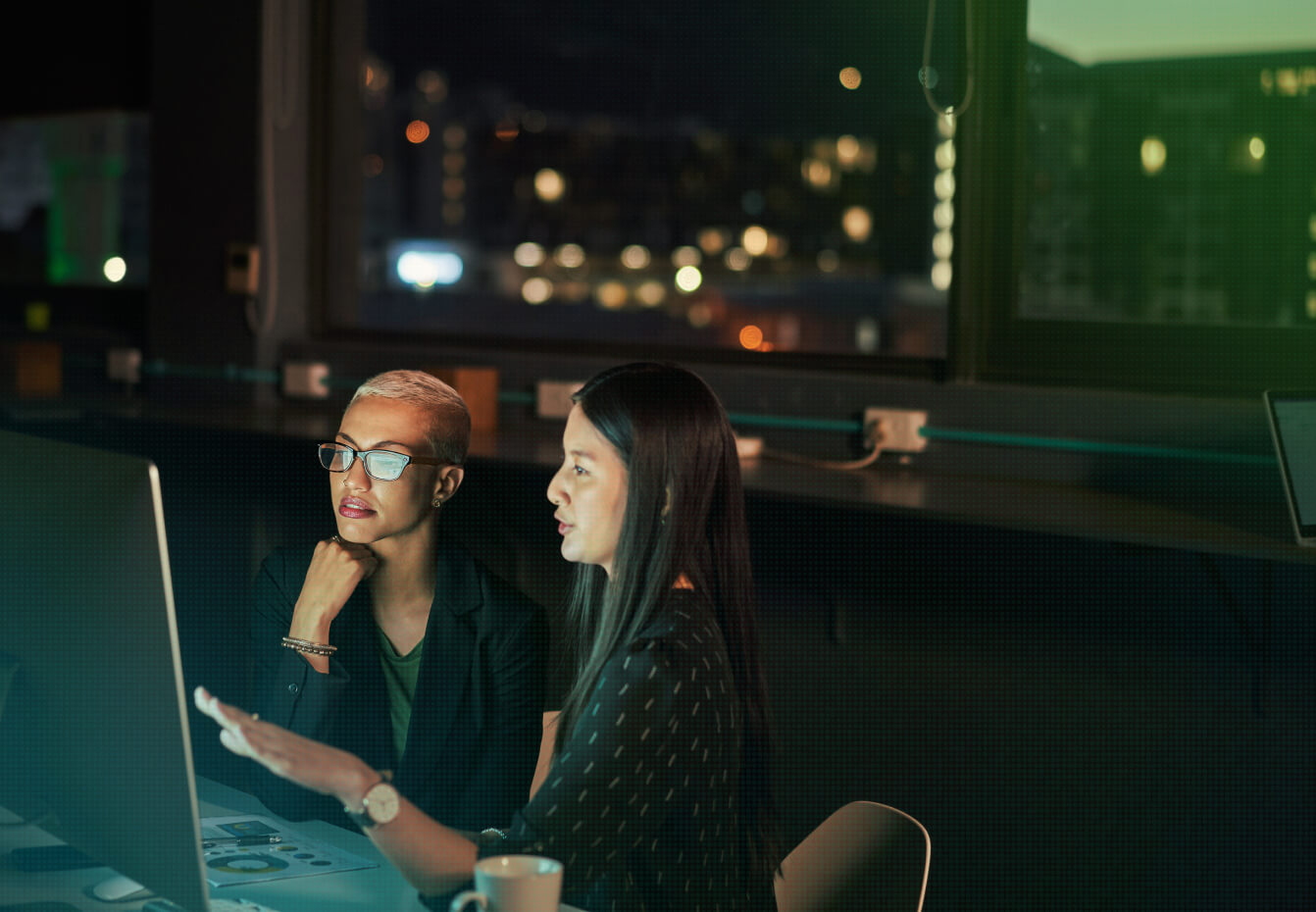 Two women in glasses collaborate at a laptop in a darkened office with a city skyline visible through the window at night.