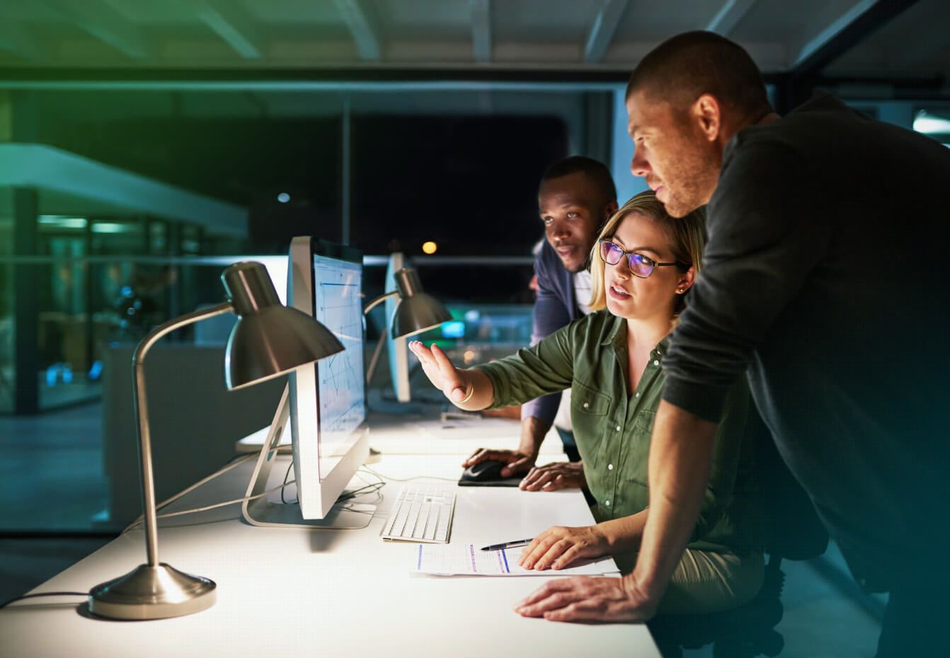 Three colleagues collaborate around a computer monitor in a dimly lit office, analyzing data on screen during a late work