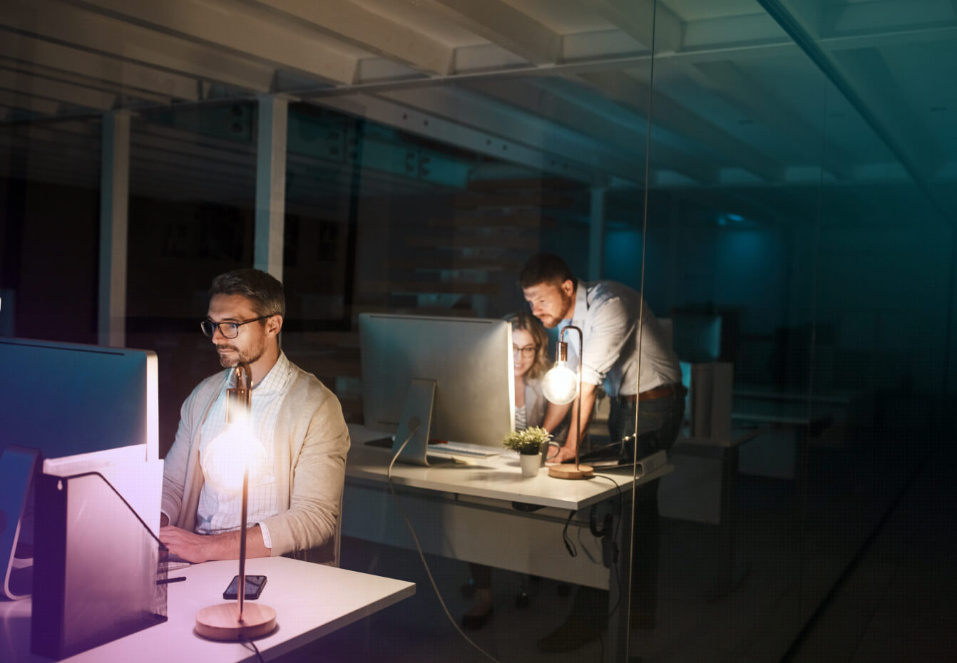 Three colleagues work late at computer workstations in a dimly lit office, illuminated by desk lamps and monitor glow.