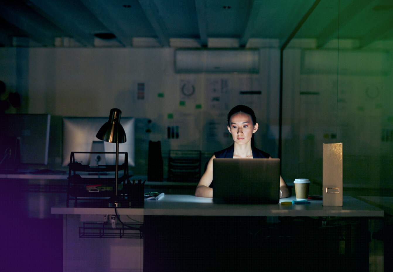 Woman working late on a laptop at her desk in a dimly lit office with a desk lamp and coffee cup nearby.