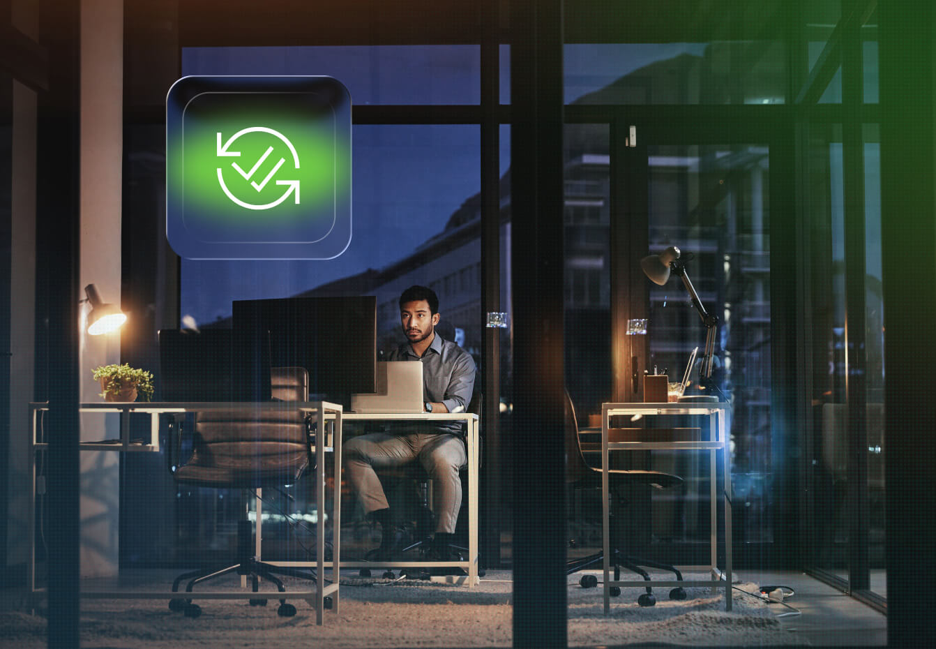 Man working late at a modern office desk with dual monitors, illuminated by lamps, beside a green compliance checkmark icon.