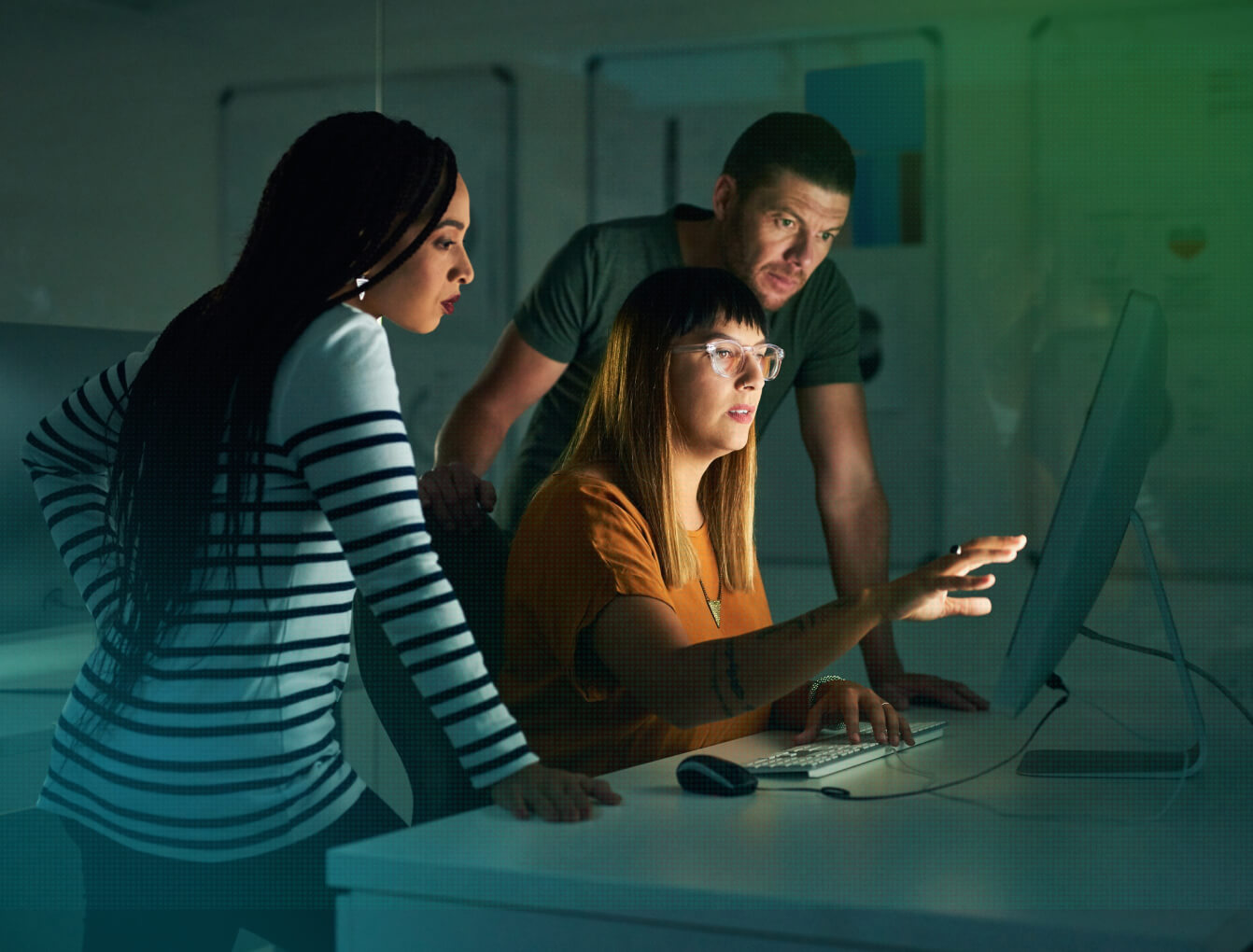 Fortreum-Use-Cases-How-XRAMP Three colleagues collaborate around a computer monitor in a dimly lit office, analyzing data together.