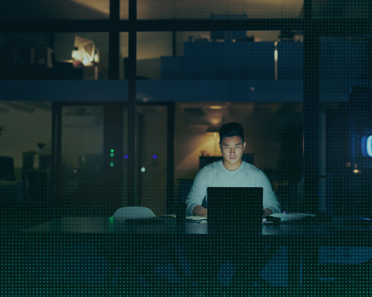 Man working on laptop in dimly lit office at night with dark teal overlay and dotted pattern effect.