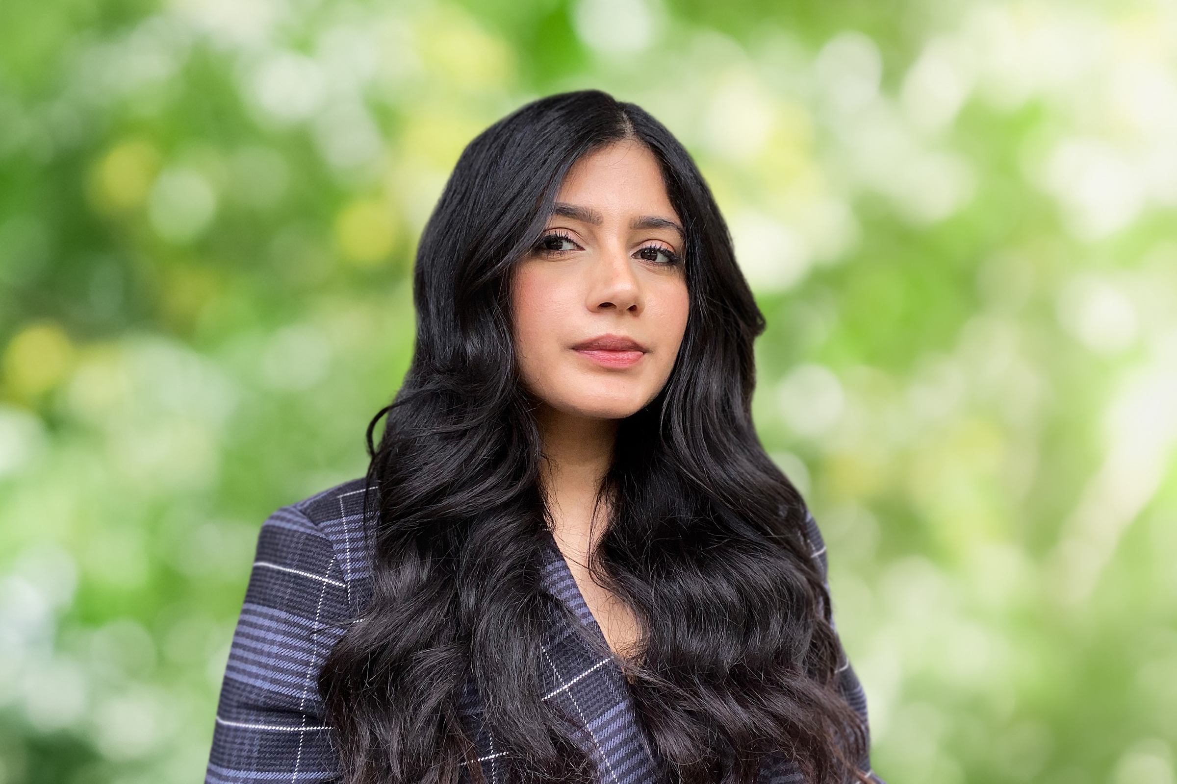 Woman with long wavy dark hair wearing a checked blazer, posed against a blurred green foliage background.