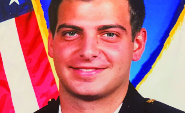 Smiling young man in a dark uniform posed in front of an American flag for an official portrait.
